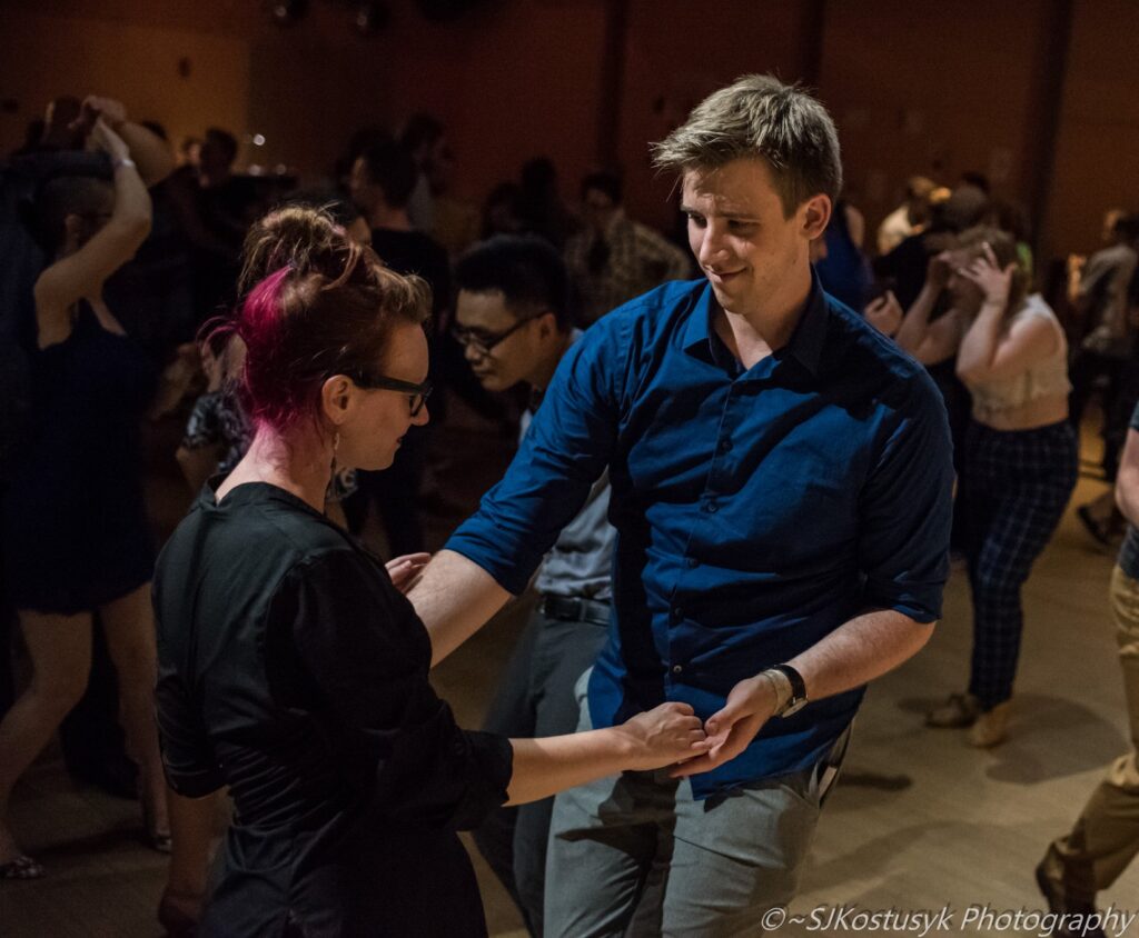 A person with pink hair and glasses dances with a very tall person in a blue shirt. Copyright SJKostusyk Photography