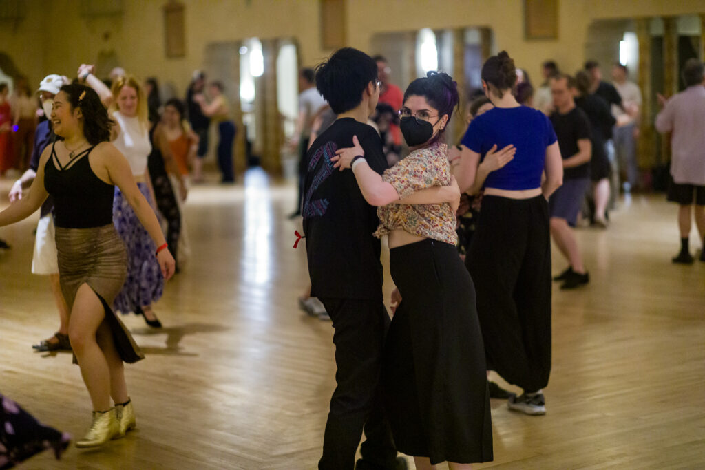 A pair of dancers in a ballroom, one in a face mask and glasses staring at the camera